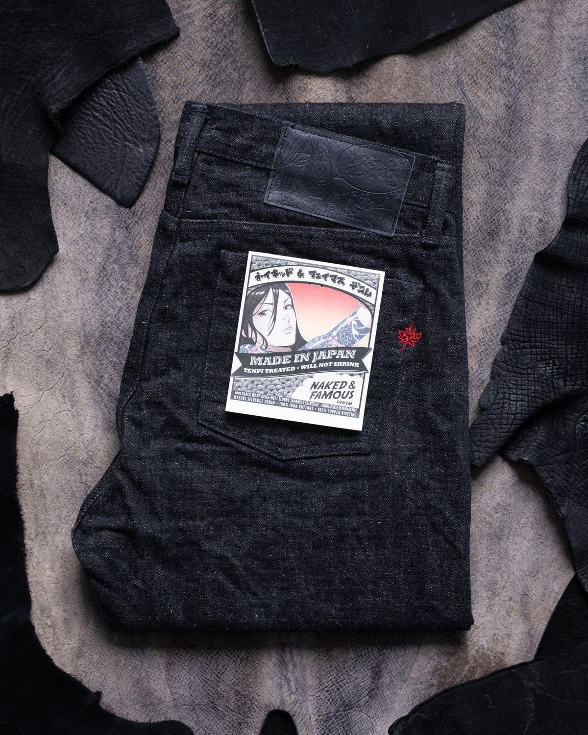 A folded pair of dark indigo denim jeans with a vintage-style back pocket patch and a small red embroidered logo, displayed on a textured gray hide background surrounded by black leather.